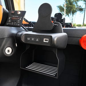 Op de afbeelding: Een zwarte dashboardconsole voor voertuigen met een centrale bekerhouder met een geribbelde bodem, een USB-poort en een bedieningspaneel met iconen voor ontwaseming, ventilator en alarmlichten. Een aparte muziekbediening is links gemonteerd.