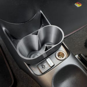 May include: A gray, 3D-printed car cup holder with two circular compartments. The cup holder is installed in a car's center console, next to a power outlet and AUX input. The console is black.