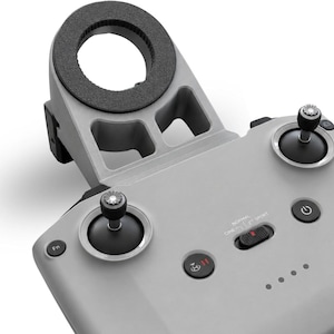 Può includere: Un telecomando per drone grigio chiaro dotato di due joystick neri, un pulsante di accensione, un selettore di modalità di volo con le diciture Cine, Normal e Sport, un tasto pausa con icona di atterraggio e un attacco circolare nero.