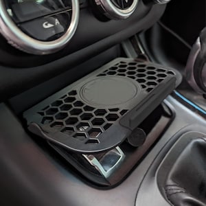 Könnte beinhalten: Ein schwarzer Autotelefonhalter mit Wabenmuster. Der Halter ist für die Mittelkonsole eines Autos konzipiert und verfügt über einen kreisförmigen Bereich zum kabellosen Laden. Der Halter besteht aus Kunststoff und hat ein schlankes, modernes Aussehen.