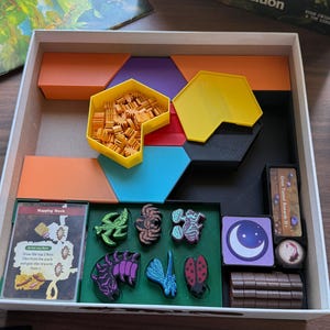 May include: A board game box containing various components including orange, purple, and blue hexagonal tiles, a yellow tray filled with small orange ant tokens, a deck of cards labeled Napping Nook, and several colorful insect-shaped game pieces.