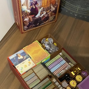 Könnte beinhalten: Ein Brettspielkasten mit dem Titel "Ada's Dream" mit einer Illustration einer Frau in einem lila Kleid. Im Inneren enthält eine rote Box Spielkomponenten: Karten, Spielsteine und Spielbretter, organisiert in Fächern.