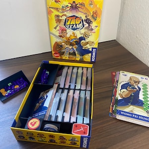 Könnte beinhalten: Eine Spielbox mit dem Titel "TAG TEAM" und Illustrationen von Zeichentrickfiguren. Die Box ist geöffnet und zeigt Spielkomponenten wie Karten, Spielsteine und ein Spielbrett. Das Spiel ist für zwei Spieler konzipiert.