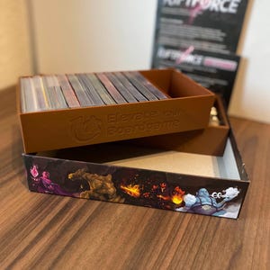 Könnte beinhalten: Eine braune Brettspielbox mit dem Text "Elevate your Boardgame" und einem Stapel Karten darin. Die Box steht auf einer Holzoberfläche, mit einem "Riftforce" Spielanleitung im Hintergrund. Die Box hat eine farbenfrohe Illustration an der Seite.