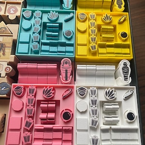 Könnte beinhalten: Ein Brettspiel-Organizer mit vier farbigen Tabletts: Türkis, Gelb, Rosa und Weiß. Jedes Tablett enthält verschiedene Spielsteine, darunter rechteckige Blöcke, kreisförmige Token und stilisierte Symbole. Die Teile sind ordentlich in den Tabletts angeordnet.