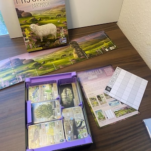 Peut inclure: Présentation du jeu de société "Mischwald Dartmoor", avec une boîte illustrée d'un mouton dans un paysage. Le jeu comprend des cartes, des plateaux et un compartiment violet. Le thème du jeu semble être la nature et le paysage.