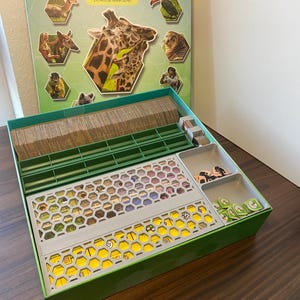 Può includere: Una scatola di gioco da tavolo intitolata "Arten Garten" con illustrazioni di animali. La scatola è aperta, rivelando componenti del gioco tra cui carte, gettoni e un tabellone con un design a nido d'ape. La scatola è verde e il tabellone è grigio e giallo.