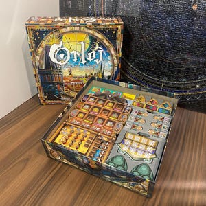 Puede incluir: Se muestra una caja de juego de mesa y su contenido. La caja presenta un diseño colorido con el título "Orloj" y el texto "The Prague Astronomical Clock". Los componentes del juego están organizados dentro de la caja abierta, mostrando varias piezas.