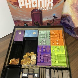 Op de afbeelding: Een geopende bordspeldoos met de titel "DAS PHÖNIX PROJEKT" in oranje en wit. Binnenin bevinden zich diverse spelonderdelen in zwart, paars, oranje en groen. De spelcomponenten omvatten fiches, kaarten en andere elementen.