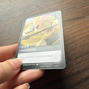 May include: A rectangular trading card titled Sand Soldier, featuring a golden armored figure in a desert landscape. The card displays the text Token Unit, Shurima, and the flavor text They do not rest. They do not hesitate. And they do wield.