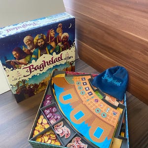 Könnte beinhalten: Brettspiel mit dem Titel "Baghdad" mit einer Box mit Illustrationen von Menschen und Kamelen. Die offene Spielbox zeigt Spielsteine, Karten und einen blauen Samtbeutel. Die Spielsteine umfassen Münzen und verschiedene Spielmarken.
