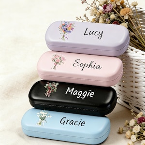 Op de afbeelding: Vier gepersonaliseerde brillenkokers in pastelkleuren: lavendel, roze, zwart en lichtblauw. Elke koker heeft een bloemenontwerp en een naam: Lucy, Sophia, Maggie en Gracie. Een witte mand en droogbloemen staan op de achtergrond.
