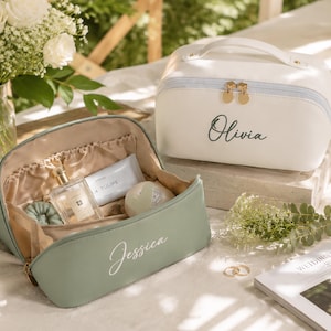 Può includere: Due beauty case rettangolari con manici superiori e cerniere dorate. Una borsa è bianca con il nome Olivia stampato in nero, l'altra è verde salvia con il nome Jessica stampato in bianco, con un interno aperto foderato in beige.