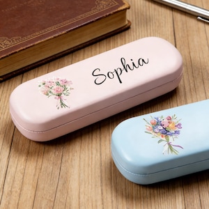 Op de afbeelding: Twee brillenkokers, één roze en één lichtblauw, met bloemboeketontwerpen. De roze koker heeft de naam "Sophia" erop gedrukt. Een bruin boek en een pen zijn ook zichtbaar op het houten oppervlak.