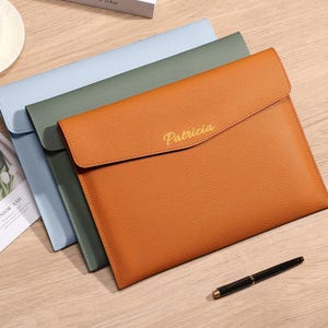 May include: Three leather document holders in shades of blue, green, and orange. The orange holder has the name "Patricia" in gold lettering. A black pen with gold accents rests on the wooden surface.