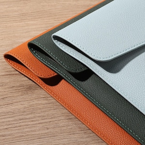 May include: Three leather envelope-style cases in orange, dark green, and light blue. Each case has a flap closure and is stitched with a contrasting thread. The cases are arranged diagonally, showcasing their textured surfaces.