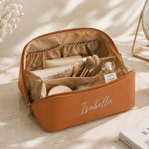 May include: A tan-colored cosmetic bag with a zipper closure and the name Isabella embroidered in white script on the front. The interior is lined with beige fabric and features multiple compartments containing various makeup brushes and bottles.