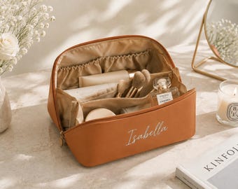 May include: A tan-colored cosmetic bag with a zipper closure and the name Isabella embroidered in white script on the front. The interior is lined with beige fabric and features multiple compartments containing various makeup brushes and bottles.