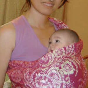 May include: A baby carrier in a vibrant pink hue, adorned with a white floral pattern. The carrier is designed to hold an infant, offering a comfortable and secure way to carry a baby. The carrier is made of soft fabric.