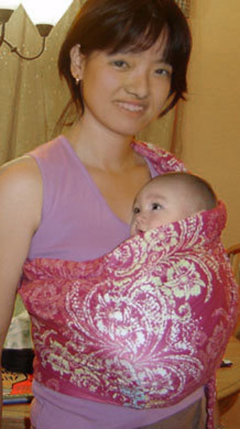 May include: A baby carrier in a vibrant pink hue, adorned with a white floral pattern. The carrier is designed to hold an infant, offering a comfortable and secure way to carry a baby. The carrier is made of soft fabric.