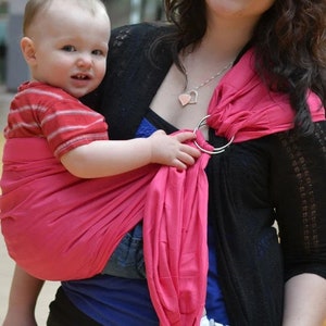May include: A fuchsia baby carrier sling with silver rings. The sling is wrapped around a baby wearing a red and white striped shirt and blue jeans. The text "Fushcia" is at the bottom of the image.