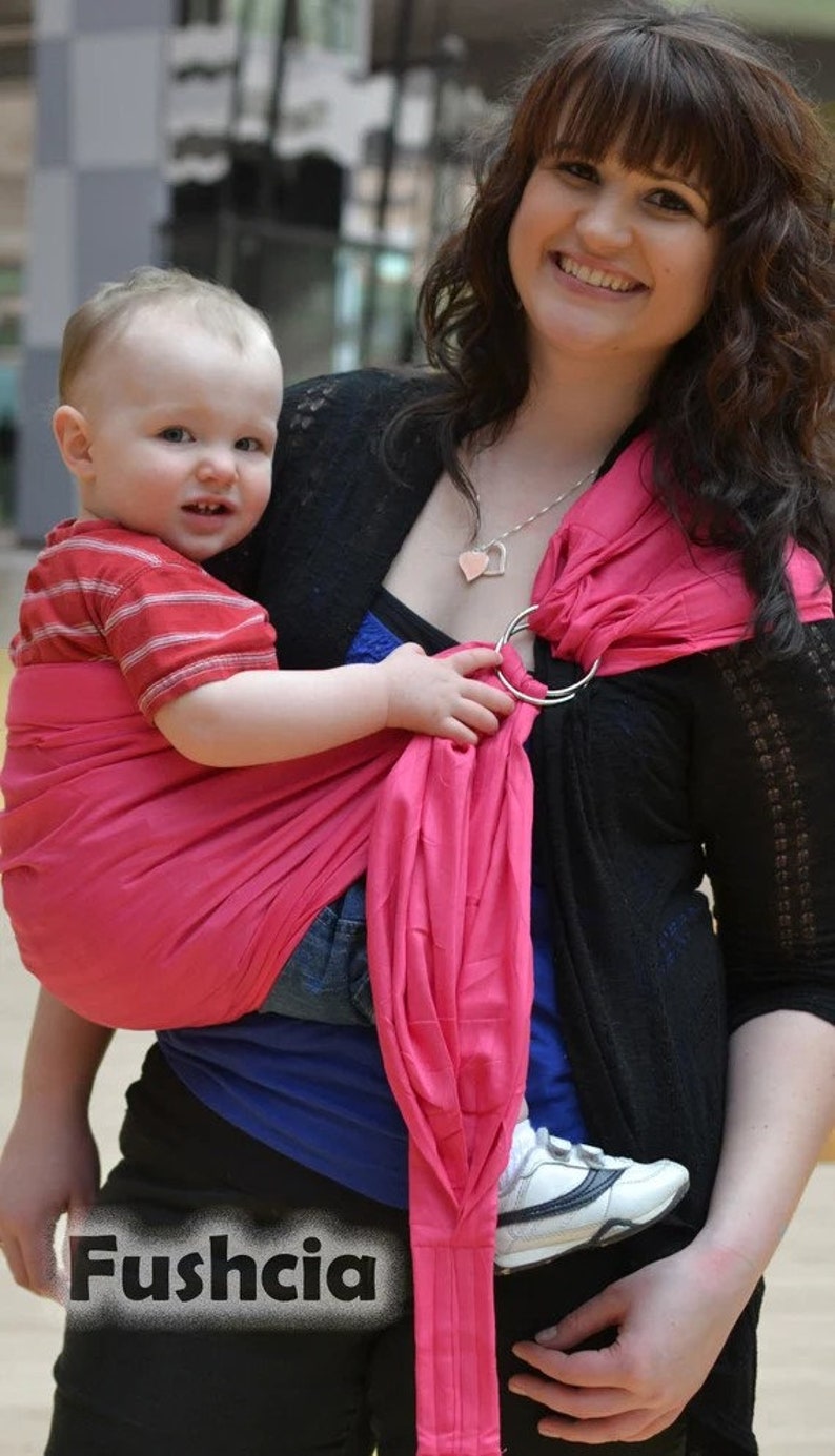 May include: A fuchsia baby carrier sling with silver rings. The sling is wrapped around a baby wearing a red and white striped shirt and blue jeans. The text "Fushcia" is at the bottom of the image.
