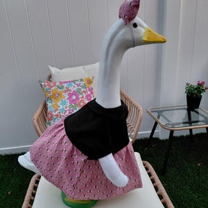 May include: A whimsical goose statue dressed in a black top and a pink skirt adorned with a bee pattern. The goose wears a matching pink bow and sits on a chair. The statue has a white body, yellow beak, and is a unique garden decoration.