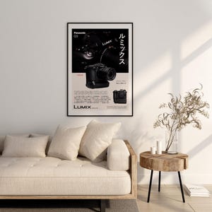 Puede incluir: Un póster enmarcado con una cámara negra Panasonic G9 con la marca LUMIX. El póster tiene texto japonés y un fondo beige. El póster está colgado sobre un sofá beige y una mesa auxiliar de madera.