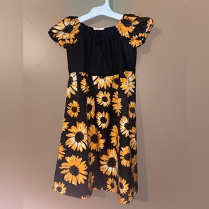 May include: A black dress with a sunflower print. The dress has a black bodice and short sleeves with a sunflower pattern. The skirt of the dress is covered in large yellow and white sunflowers on a black background.