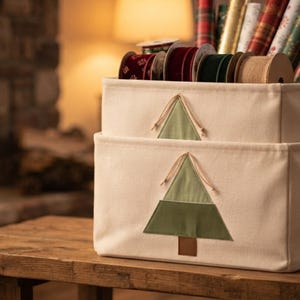 May include: Two stacked off-white rectangular fabric storage bins, each featuring a centered green fabric Christmas tree appliqué with a brown trunk. The bins are filled with various rolls of colorful wrapping paper and spools of ribbon.