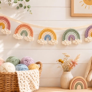 May include: A decorative garland featuring five crocheted rainbow ornaments in various pastel colors, each with white cloud accents at the base. The rainbows are suspended from a cream-colored fabric ribbon against a white horizontal paneled wall.