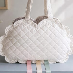 May include: A cream-colored quilted tote bag with a scalloped edge and diamond-stitched pattern. The bag features two top handles and three hanging ribbons in soft pink, beige, and sage green extending from the bottom edge of the bag.