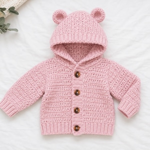 May include: A pale pink, crocheted baby hoodie with bear ears on the hood. The button-up cardigan has long sleeves and brown buttons. The garment is displayed on a white surface.