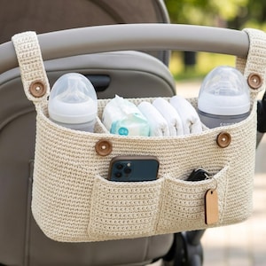 May include: A beige crochet stroller organizer featuring two main compartments, two front pockets, and two side straps with wooden buttons. The organizer holds two baby bottles, a pack of wipes, a smartphone, and a set of keys with a wooden tag.