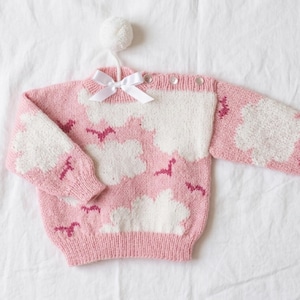 May include: A long-sleeve knit sweater in light pink featuring a white cloud pattern and small dark pink bird motifs. The sweater has a white satin bow at the neckline, a white pom-pom detail, and three buttons along the left shoulder seam.