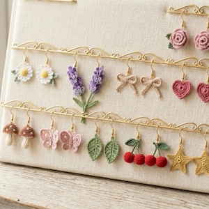 May include: A beige fabric earring display stand featuring multiple pairs of crocheted earrings in various shapes including flowers, lavender sprigs, bows, hearts, mushrooms, butterflies, leaves, cherries, and stars, all hanging from gold metal bars.