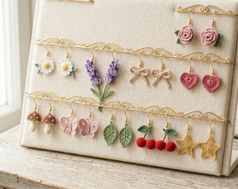 May include: A beige fabric earring display stand featuring multiple pairs of crocheted earrings in various shapes including flowers, lavender sprigs, bows, hearts, mushrooms, butterflies, leaves, cherries, and stars, all hanging from gold metal bars.