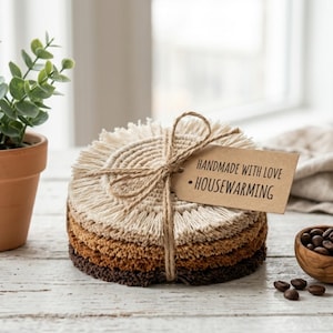 May include: A stack of four round, fringed coasters in gradient shades of cream, tan, light brown, and dark brown. The coasters are tied together with jute twine and feature a brown paper tag with the text HANDMADE WITH LOVE • HOUSEWARMING.