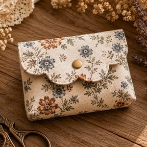 May include: A small rectangular fabric pouch with a scalloped flap closure secured by a brass-colored snap button. The pouch features an off-white base color with a repeating floral pattern in shades of muted blue, rust orange, and sage green.