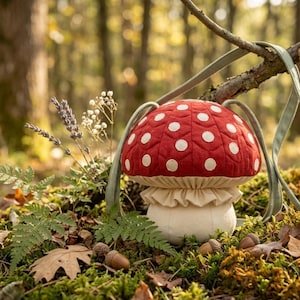May include: A mushroom-shaped crossbody bag featuring a quilted red cap with white polka dots, a cream-colored ruffled collar, and a solid cream stem. The bag includes a long, light green strap attached to the top of the mushroom cap.