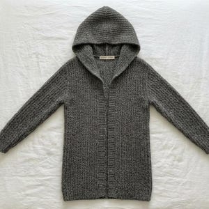 May include: A charcoal gray open-front hooded cardigan featuring a thick ribbed knit texture. The garment has long sleeves and a hood, with a small white label visible at the inner neckline. The knit pattern is consistent throughout the piece.