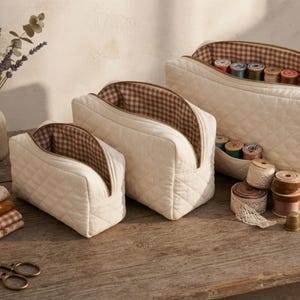 Può includere: Tre pochette trapuntate color crema di varie dimensioni con fodera interna a quadretti vichy marroni e bianchi e cerniere color oro. Le pochette sono aperte e rivelano rocchetti di filo all'interno, con altri articoli da cucito.