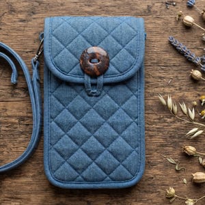 May include: A blue quilted fabric phone pouch with a long matching shoulder strap. The rectangular pouch features a diamond-stitched pattern and a flap closure secured by a single large, dark brown, circular wooden button with a central hole.