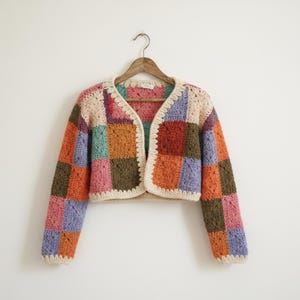 May include: A colorful, patchwork crochet cardigan with a cream-colored trim and a wooden hanger. The cardigan features squares of orange, pink, purple, green, and cream-colored yarn. The sleeves are long and the cardigan is open in the front.
