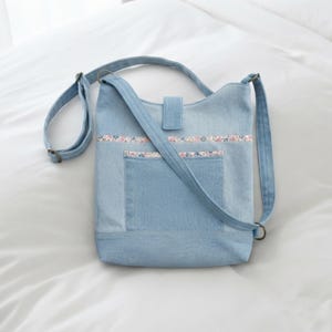 Könnte beinhalten: Eine hellblaue Umhängetasche aus Denim mit verstellbarem Schulterriemen, einer zentralen Außentasche und einem Klappenverschluss. Die Tasche ist mit einem horizontalen Streifen aus geblümtem Stoff an der Oberseite und am Taschenrand verziert.