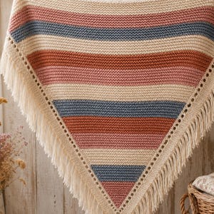 Può includere: Uno scialle triangolare in maglia con strisce orizzontali color crema, rosa antico e blu ardesia. Il capo presenta un bordo decorativo con occhielli lungo i due lati inclinati ed è rifinito con lunghe frange color crema sui lati inferiori.