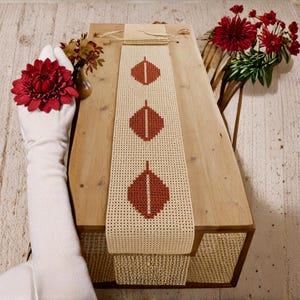 May include: A long, rectangular, cream-coloured crochet table runner featuring three vertically aligned, rust-coloured leaf motifs created with a cross-stitch pattern. The runner has a textured, grid-like weave and is finished with a simple knotted fringe.