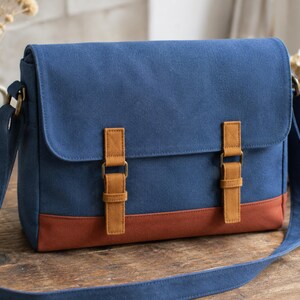 May include: A blue canvas messenger bag featuring a large front flap, two tan faux leather buckle straps, and a solid brown base panel. The bag includes an adjustable blue shoulder strap with antique brass-toned metal hardware and side rings.