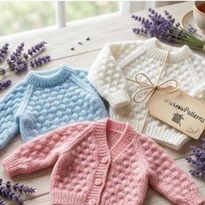 Könnte beinhalten: Drei handgestrickte Baby-Pullover in Pastellfarben: Hellblau, Weiß und Rosa. Die Pullover haben ein strukturiertes Muster und werden mit Lavendelzweigen und einem Etikett mit der Aufschrift "littleewPatterns" präsentiert.