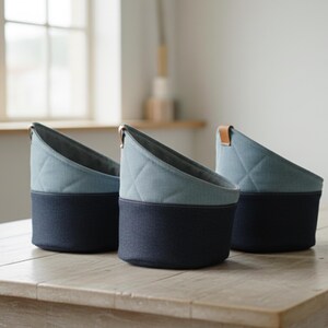 Könnte beinhalten: Drei zweifarbige Stoff-Aufbewahrungskörbe. Der untere Teil ist marineblau und der obere Teil hellblau mit einem Steppmuster. Sie stehen auf einem hellen Holztisch.